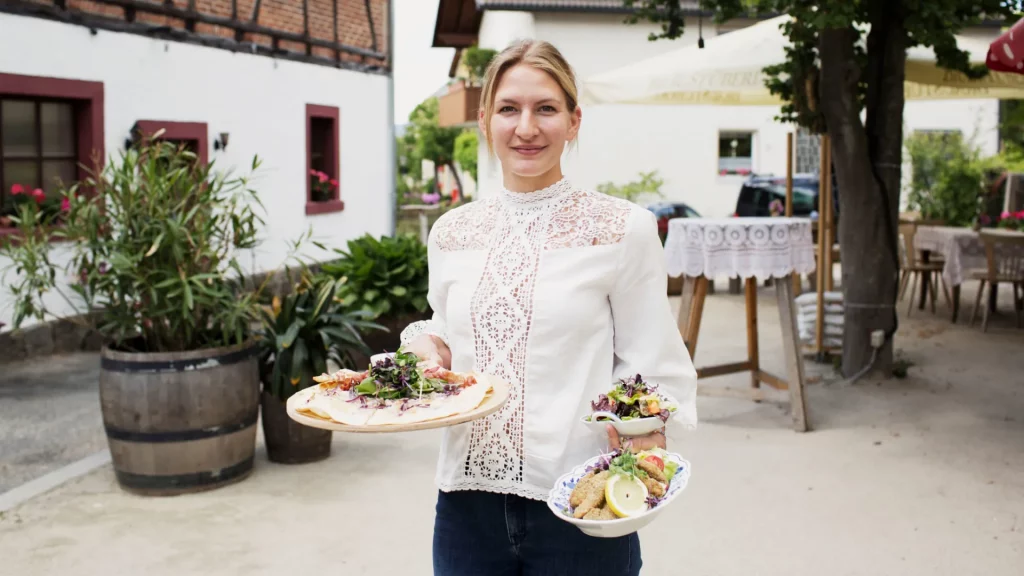 Eine Bedienung hält in der rechten Hand eine Essensplatte, in der rechten trägt sie zwei Nebenspeisen. Im Hintergrund ist ein Biergarten erkennbar.