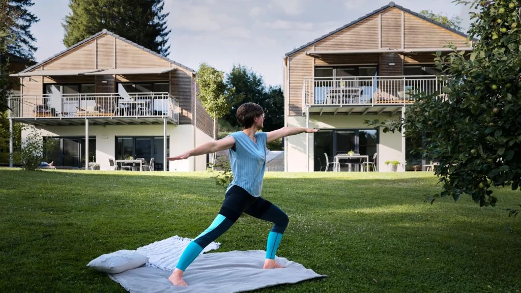Eine Frau auf einer Wiese macht Yoga Übungen. Im Hintergrund sieht man zwei identische Ferienhäuser.