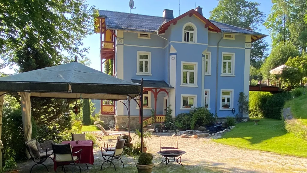Ein altes Haus frisch saniert von außen fotografiert. Im Vordergrund sieht man einen Pavillon, rechts einen Aufgang im Garten.