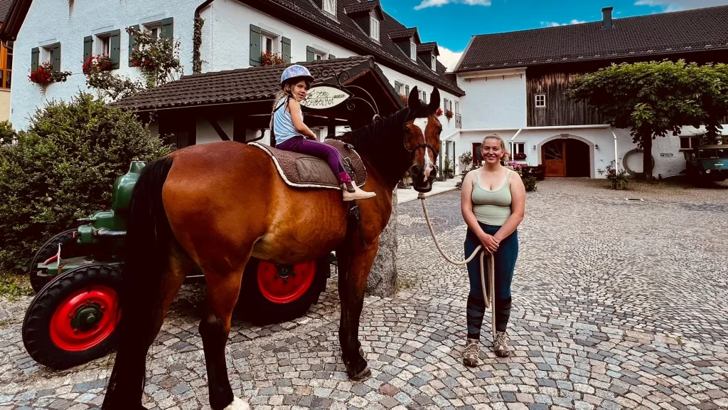 Vor einem Ferienhof steht eine Frau, neben ihr ein Kind das auf einem Pferd reitet.